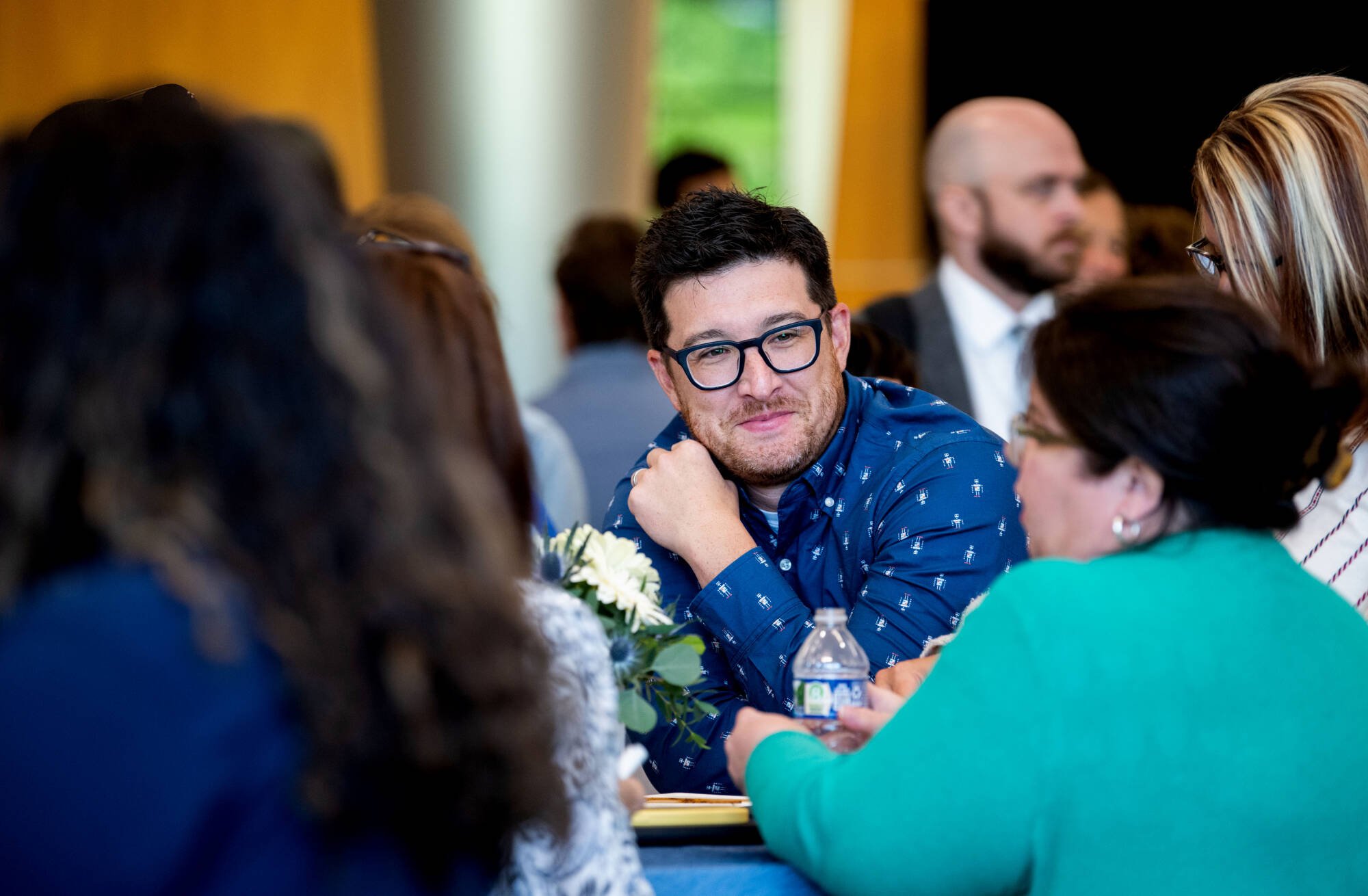 A man in a blue patterned shirt with glasses converses at a table with a woman in a green sweater.